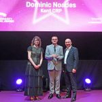 Dominic Noades on stage at the Community Rail Awards 2026. He is receiving his award from the hosts of the evening.