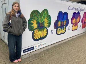 A person stand beside a vinyl on the side of a byilding featuring flowers and "Garden of England" at the top and "KENT" in individual letters between the flowers.
