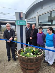 Chris cuts a ribbon above a planter on the platform of a station while people look on