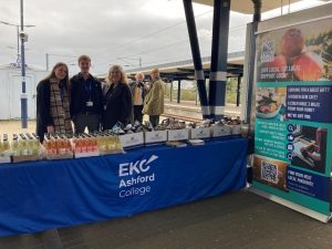 People standing at a refreshments stall with EKC Ashford College logo and a roller banner promoting the college