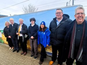 A group of people pose for a photo in front of a large vinyl displaying golden wheat fields under a blue sky