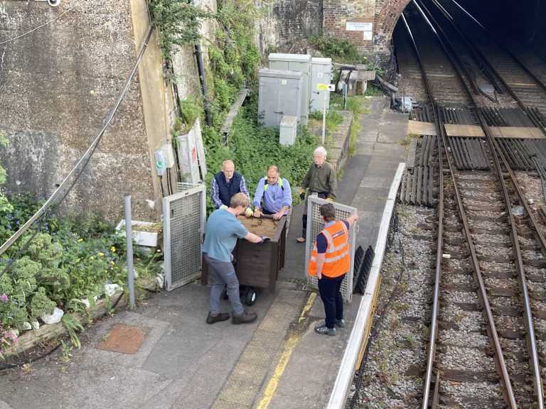 A group of people wheel a large wooden planter on a trolley onto a station platform from track level. A member of railway staff is escorting them