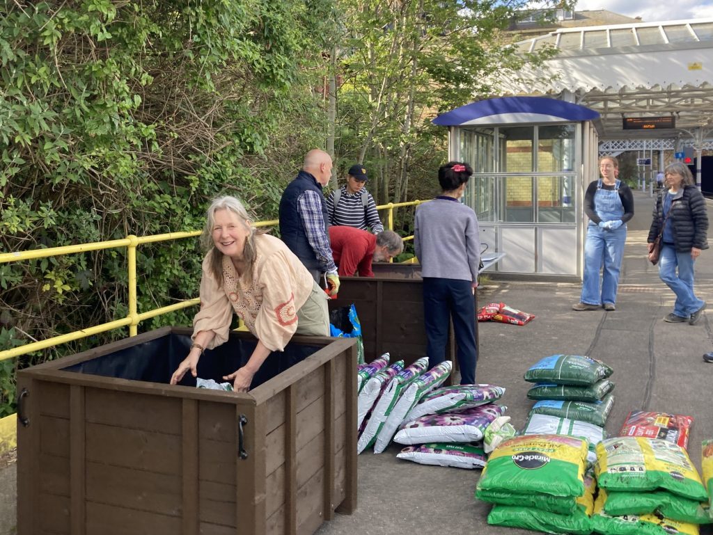 A pair of wooden planters on a station platform, bags of peat free compost, and soil. Seven people are helping to fill the planters
