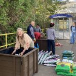 A pair of wooden planters on a station platform, bags of peat free compost, and soil. Seven people are helping to fill the planters