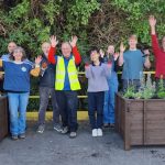 A group of 10 people wave their arms, as they celebrate completing the installation of two wooden planters filled with soil and fresh planting.