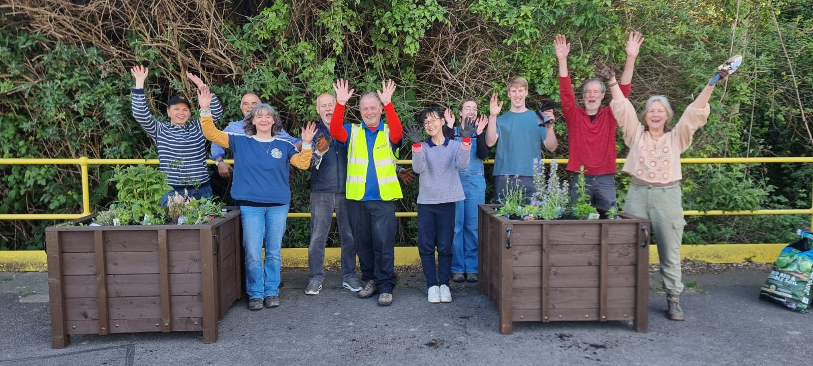 A group of 10 people wave their arms, as they celebrate completing the installation of two wooden planters filled with soil and fresh planting.