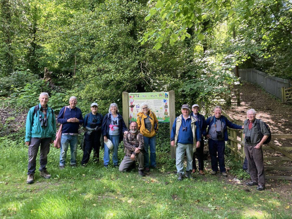 A group of people stand in a line as they pose for a photo. Behind them are tress in full green leaf and a noticeboard for Oldpark Hill nature reserve