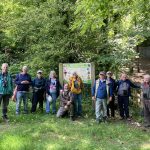 A group of people stand in a line as they pose for a photo. Behind them are tress in full green leaf and a noticeboard for Oldpark Hill nature reserve