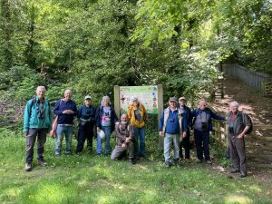 A group of people stand in a line as they pose for a photo. Behind them are tress in full green leaf and a noticeboard for Oldpark Hill nature reserve