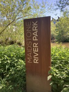 A rusted looking monolith placemaking sign for Maidstone River Park, set in a green verge at the edge of the path.