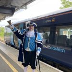 A person dressed in a dark and light blue town criers' suit rings a large bell. He is standing on a platform at a station. Behind him is a dark blue train. with ight blue doors. The colours match the town crier's uniiform.