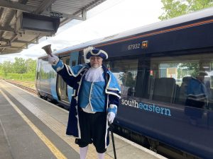 A person dressed in a dark and light blue town criers' suit rings a large bell. He is standing on a platform at a station. Behind him is a dark blue train. with ight blue doors. The colours match the town crier's uniiform.