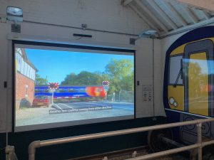 A large cinema screen displays an image of a train crossing a level crossing. On the wall to one side is a life size illustration of the front of a train.