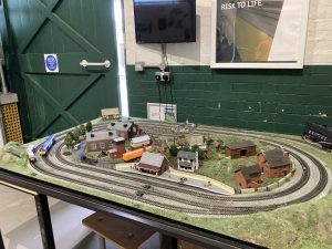 A model railway layout with track, trains and buildings. Looking closely will reveal model people engaged in poor behaviours, for example tresspassing and sitting with legs dangling over the platform's edge.