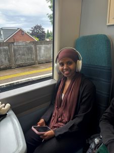 A person wearing a black jacket and trousers, with headphones on sitting onboard a train