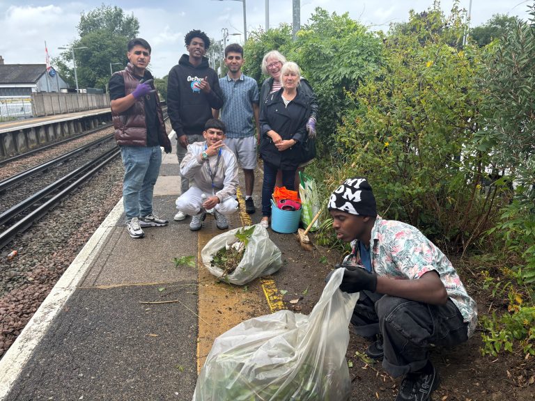 A group of people on a narrow station platform pose for a photo. There are bags of weeds that have been removed from an adjacent flower bed and a bucket of fresh plants. One person is still actively weeding whilst the photo is being taken.