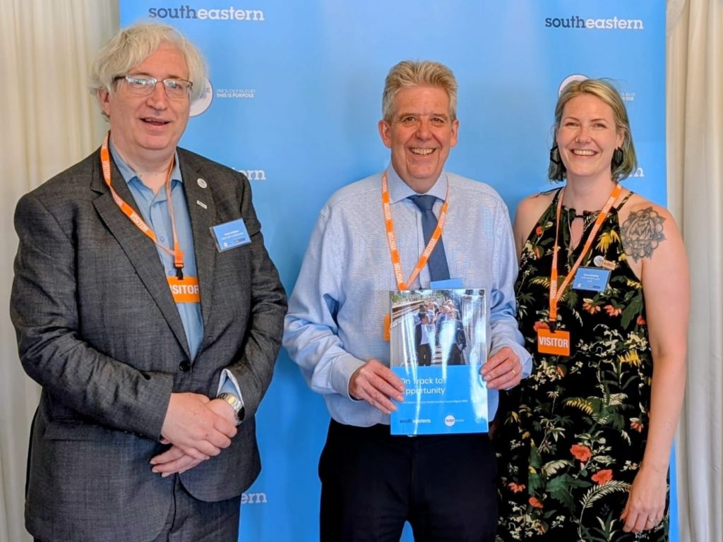 Chris, Matt and Clare pose in front of a Southeastern event roller banner for a picture. Matt is holding a copy of the "On track to opportunity" report.