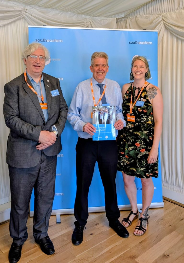 Chris, Matt and Clare pose in front of a Southeastern event roller banner for a picture. Matt is holding a copy of the "On track to opportunity" report.