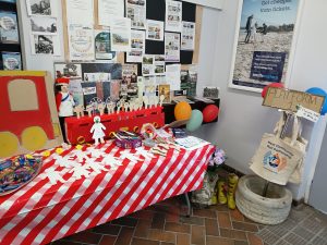 Corner of a station ticket office. A noticeboard on the wall is full of historical pictures, posters and information. A table in front holds cut out cardboard figures of people waiting for decoration. There are pens, pencils and other materials to be creative with. Hanging from a stand to one side are Kent Community Rail partnership tote bags. There is a "help yourself" tray of bagged sweets.