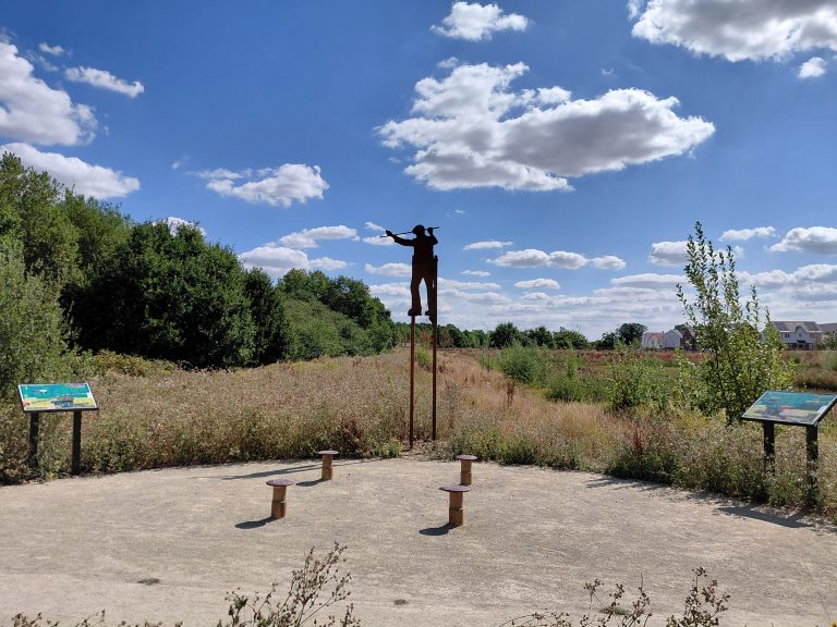 A clearing at the edge of scrubland with views to fields, houses, trees and hedgerows beyond. In the clearing are four seats fashioned from railway buffers, two information panels and a sculpture of a man on tall stilts. The sky is blue with light cloud.