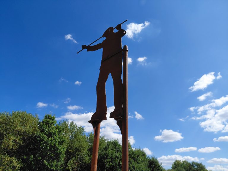 A sculpture of a man on tall stilts silhouetted against the sky and treetops. The sky is blue with light cloud.