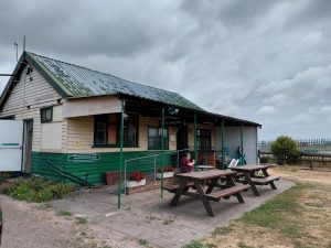 A weather-worn green and cream coloured wooden building with benches on a grassed area outside. It has a green corrugate roof.