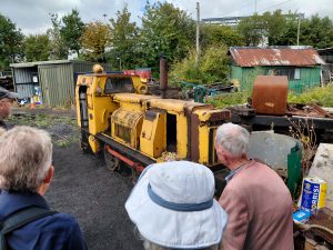 A group of people are gathered around a small rusty yellow diesel locomotive in an engine yard. There are sheds in the background.