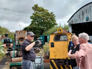 A group of people are gathered around a small rusty yellow diesel locomotive in an engine yard. There are sheds, a boiler and a steam locomotive in the background.
