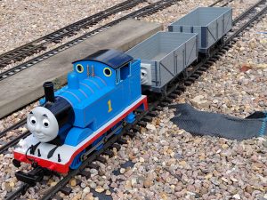 A close up of an outdoor model railway, featuring Thomas, a blue tank engine with two troublsome trucks.