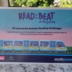 A Southeastern Summer Reading Challenge Activity pack propped up on a table onboard a train. Titled "Read to the beat of the journey" the cover shows a train full of families seated by the train windows, colourful bunting is strung along the outside of the train.