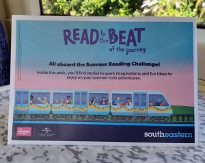 A Southeastern Summer Reading Challenge Activity pack propped up on a table onboard a train. Titled "Read to the beat of the journey" the cover shows a train full of families seated by the train windows, colourful bunting is strung along the outside of the train.