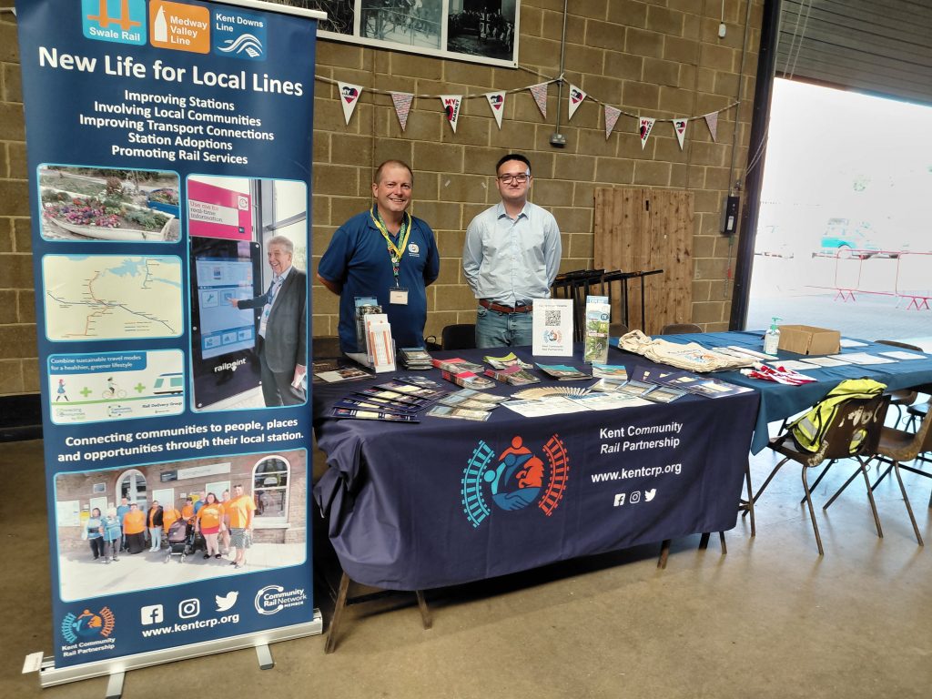 Gary and Dominic at the Kent Community Rail Partnership stand. There is a roller banner, a table full of promotional items and brochures and a second table with colouring in and activity sheets.