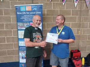Vincent and Gary are shaking hands in front of a Kent Community Rail Partnership promotional roller banner. Gary is handing Vincent a certificate of appreciation for Maidstone CAN, station adoption group.