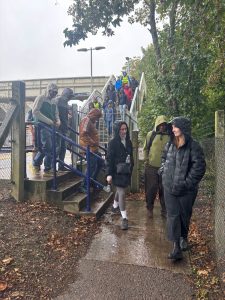 A group of over 20 people are descending from a station footbridge onto a paved path. The sky is grey, People are wearing raincoats, most with hoods up, it is raining.