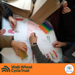 Photograph looking down at a table, childrens hands can be seen completing colouring in and activity sheets. An orange footer displays the logos of Walk Wheel Cycle Trust and Kent Community Rail Partnership.