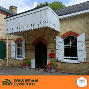 A station building. A white wooden canopy extends above the door, to either side are windows with white wooden shutters swung open to let light in. Either side of the door are low concrete planters with colourful floral displays. An orange footer displays the logos of Walk Wheel Cycle Trust and Kent Community Rail Partnership.