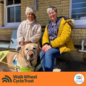 Two people seated on a bench on a station platform. In front of them is a guide doh in working harness. An orange footer displays the logos of Walk Wheel Cycle Trust and Kent Community Rail Partnership.