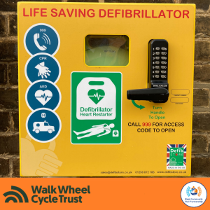 A yellow box with a pin code nadle mounted on a brick wall. "Life Saving Defibrillator" Icons indicate to call 999, perform CPR, use the Defibrillator and transfer the casualty to hospital by ambulance. An orange footer displays the logos of Walk Wheel Cycle Trust and Kent Community Rail Partnership.