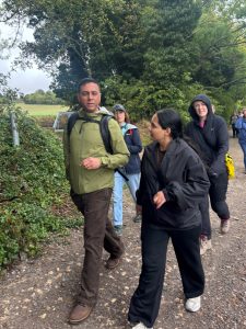 A group of people walk along a gravel path in woodland. Behind them is an open field on a hillside. The sky above is grey.