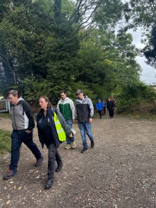 A group of people walk along a gravel path in woodland. The sky above is grey.