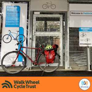 A red bicyle with panniers and a cycle helmet on the back parked outside the Paddock Wood station cycle hub An orange footer displays the logos of Walk Wheel Cycle Trust and Kent Community Rail Partnership.