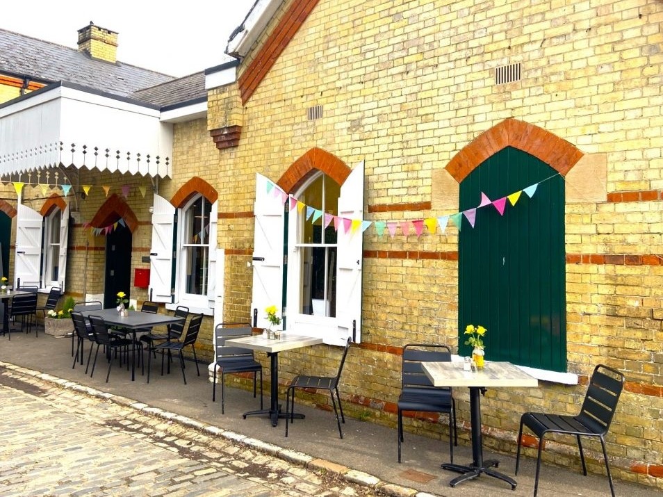 A brick built station building with shuttered windows. The outsides of the closed shutters are green, the insides of the open shutters are white. A white painted wooden canopy extendsabove the green entrance doors. There are tables and chairs outside with vases of yellow daffodils. Colourful bunting is strung between the windows and under the canopy. An orange footer displays the logos of Walk Wheel Cycle Trust and Kent Community Rail Partnership.