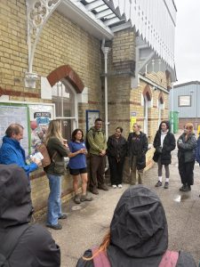 A group of people listen to Therese presenting outside a station