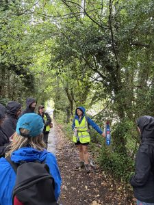 Therese talks to a group of people on a woodland path. She is pointing to trail markers on a wooden post beside the path.