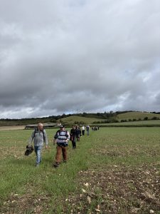A group of people walk along a path across a field. Behind them are open fields on a hillside. The sky above is grey.