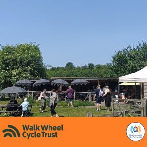 People seated at woodne benches with parasols and milling about outside a shed like building. Unde its shelter are more seats and a counter area serving drinks and snacks. An orange footer displays the logos of Walk Wheel Cycle Trust and Kent Community Rail Partnership.