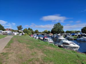 Boats moored on the river, with more out of the water in a marina on the far bank. A bridge crosses the river ahead. The sky is a bright blue with light cloud.