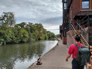 A wide river with ducks swimming towards camera. the far bank is lined with trees. The nearside path has bollards to tie up moored boats and buildings in an industrial warehouse style. Mike is walking ahead.