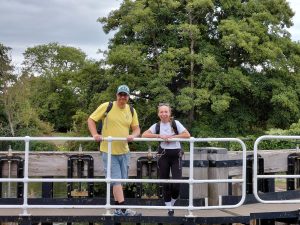 James and Roxy pose for a picture leaning on the crossing handrail of a lock gate. Trees in full leaf form the backdrop.