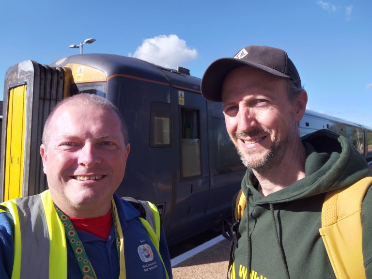 A selie of Gary and Stuart on a train platform with a train in the background. it is a bright sunny day.